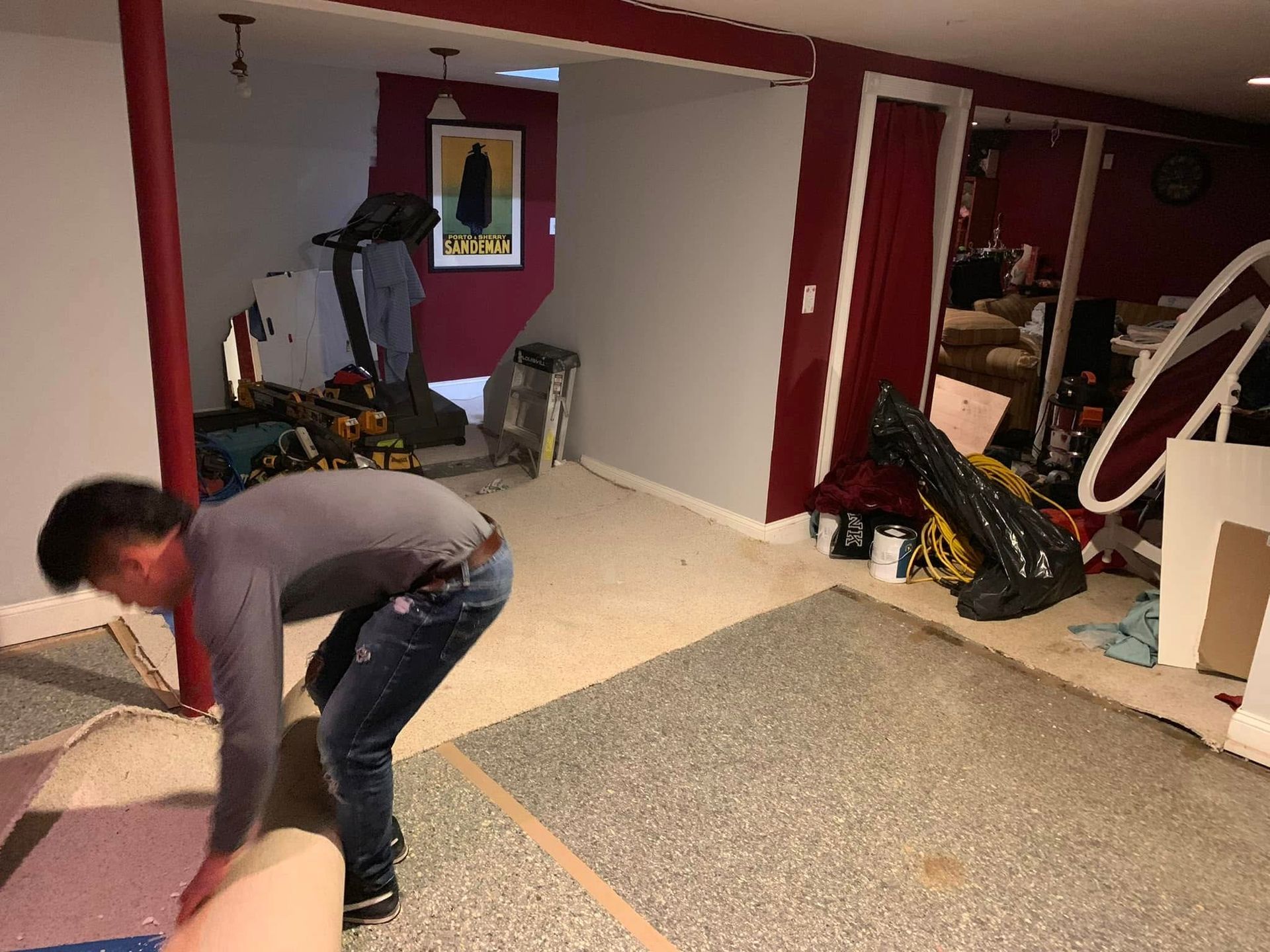 A person installing carpet in a basement with construction materials and a treadmill in the background.