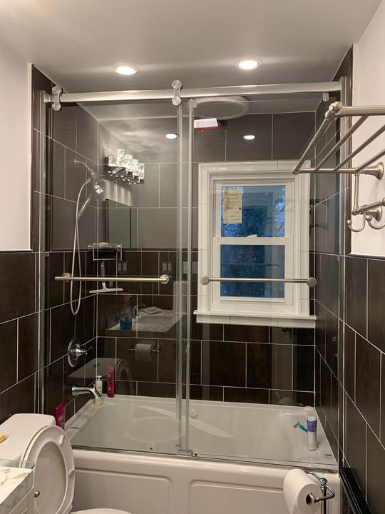 Bathroom with glass shower doors, dark tile, a window, and a bathtub.