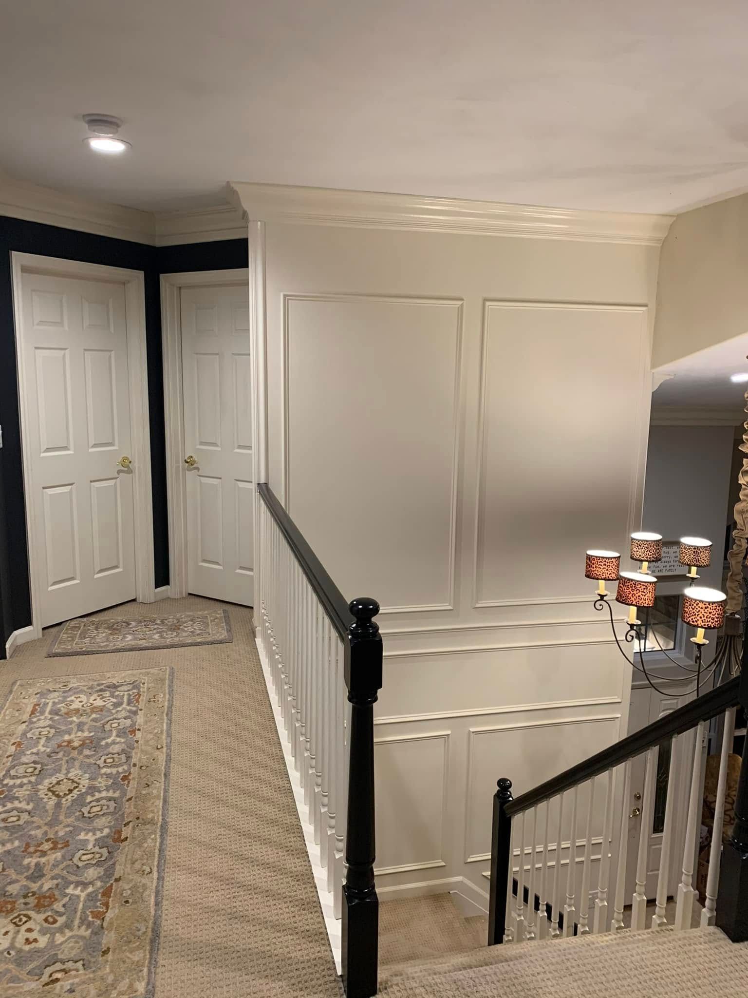 Hallway with stairs and white paneling. Black banister, beige carpet, and white doors.