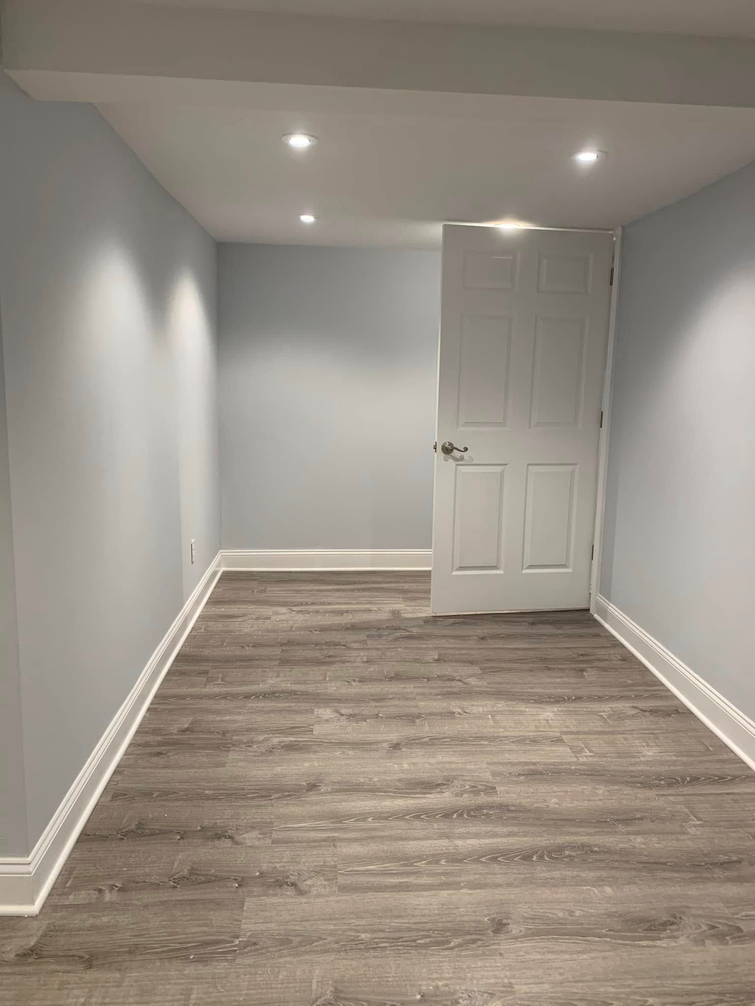 Empty room with gray walls, wood-look floor, white door and trim, recessed lighting.