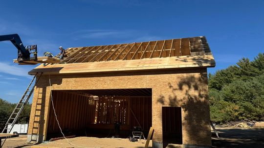 Garage under construction: Workers on roof, using a lift. Blue sky, wooden structure.