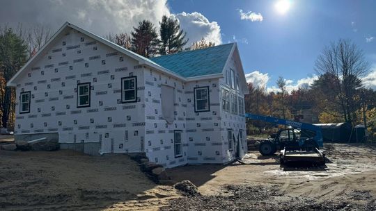 Construction of a two-story house, wrapped in protective material, under a sunny sky.
