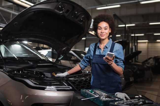 Mechanic in blue overalls standing by an open car hood, holding a phone, smiling. Tools are in front of the car in a garage.