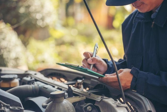Mechanic in a blue uniform inspecting a car engine, writing on a clipboard outdoors.