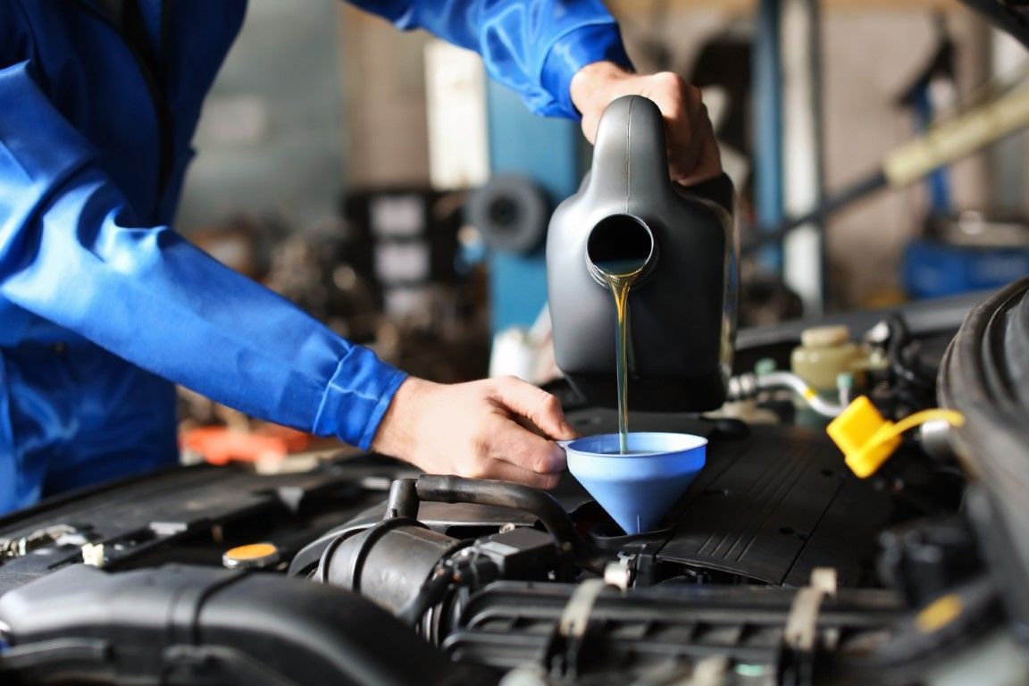 Mechanic in blue coveralls pouring oil from a black container into a car engine using a funnel. Automotive shop.