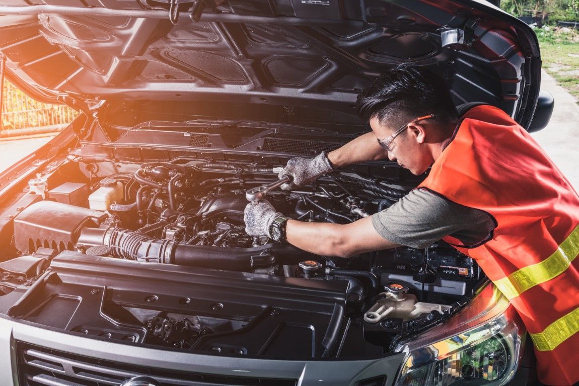 A mechanic in an orange safety vest works on a car engine with the hood open.