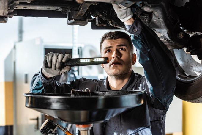 Mechanic in blue overalls examining a car's undercarriage, holding a light and oil drain pan in a garage.