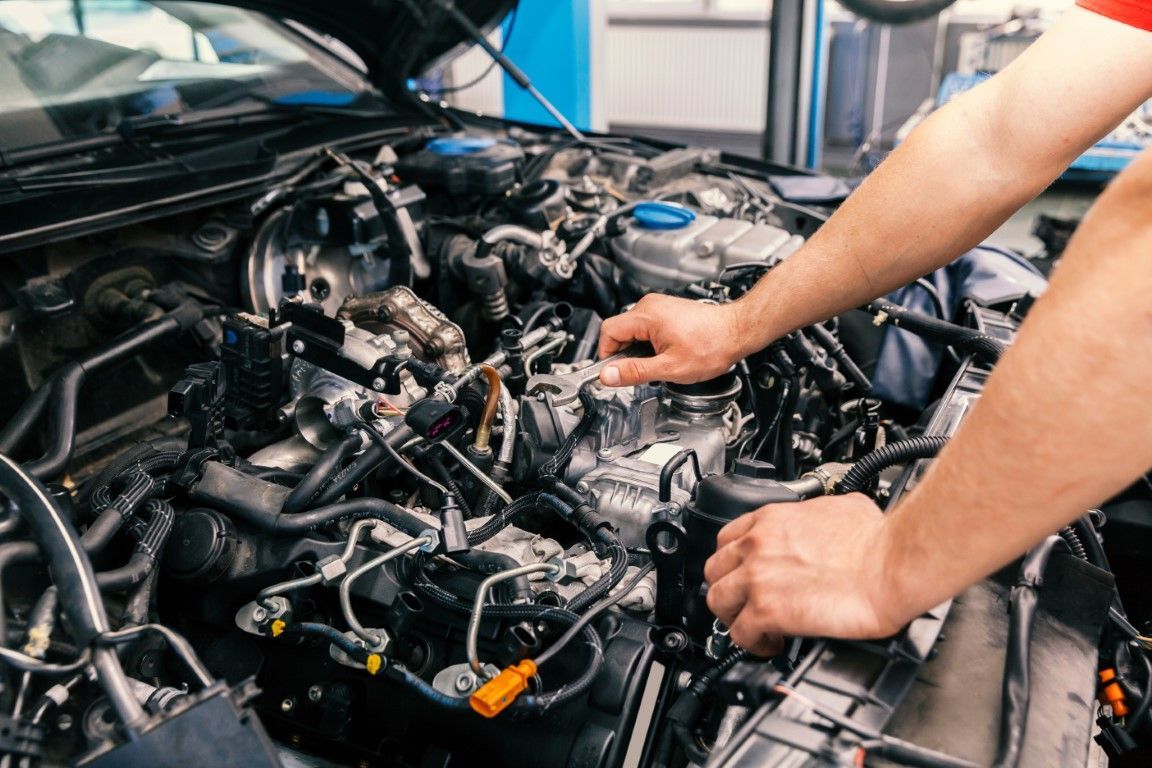 Hands working on a car engine, likely in a repair shop. The engine is black, with visible wires and tubes.