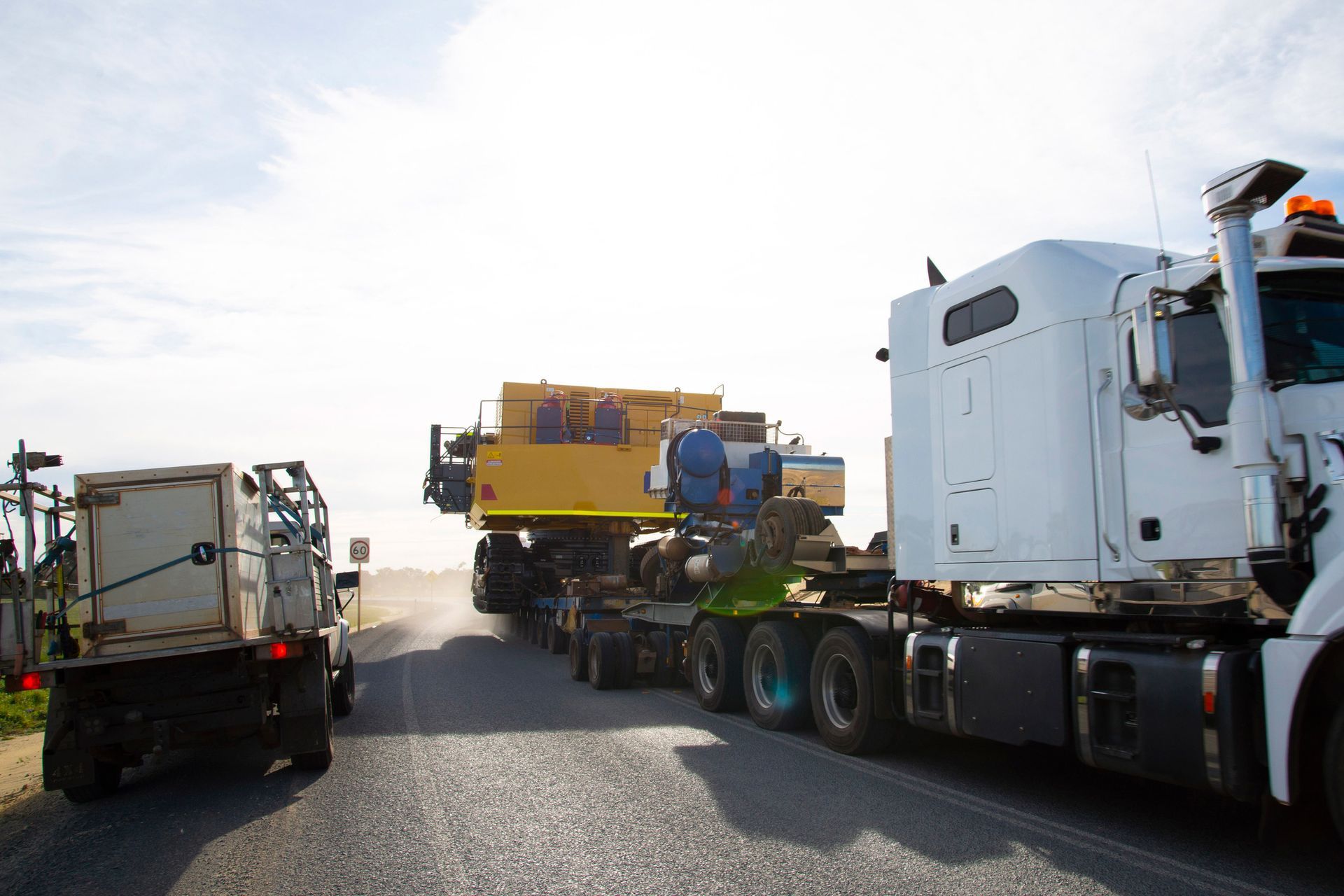 A white semi truck is carrying a yellow excavator on a trailer.
