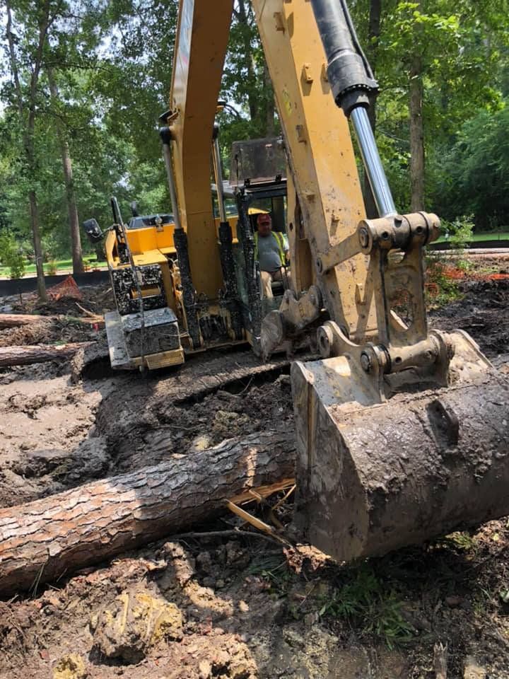 Yellow excavator removing a tree trunk from muddy ground in a wooded area.