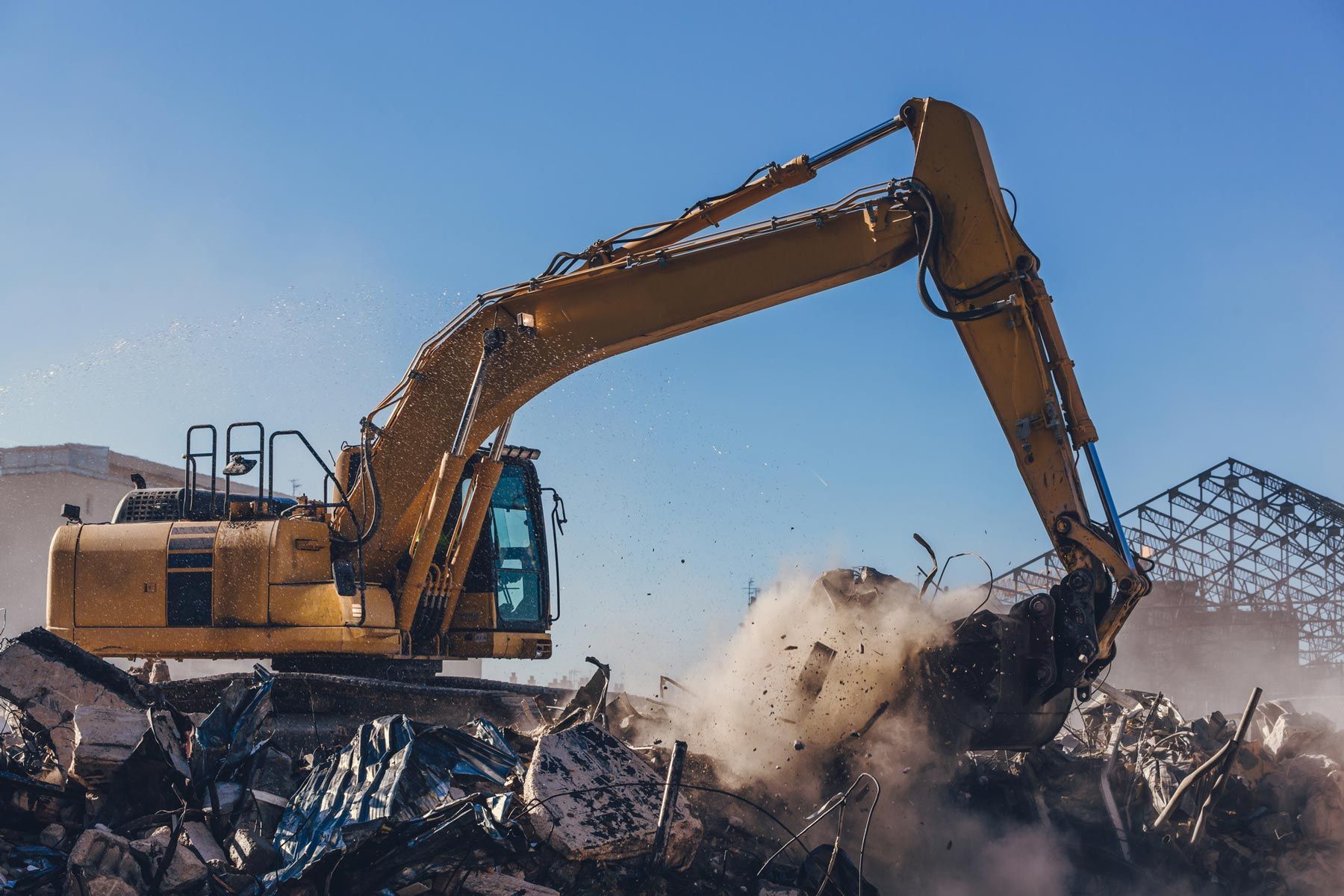 Yellow excavator demolishing a pile of debris outdoors under a clear, blue sky.