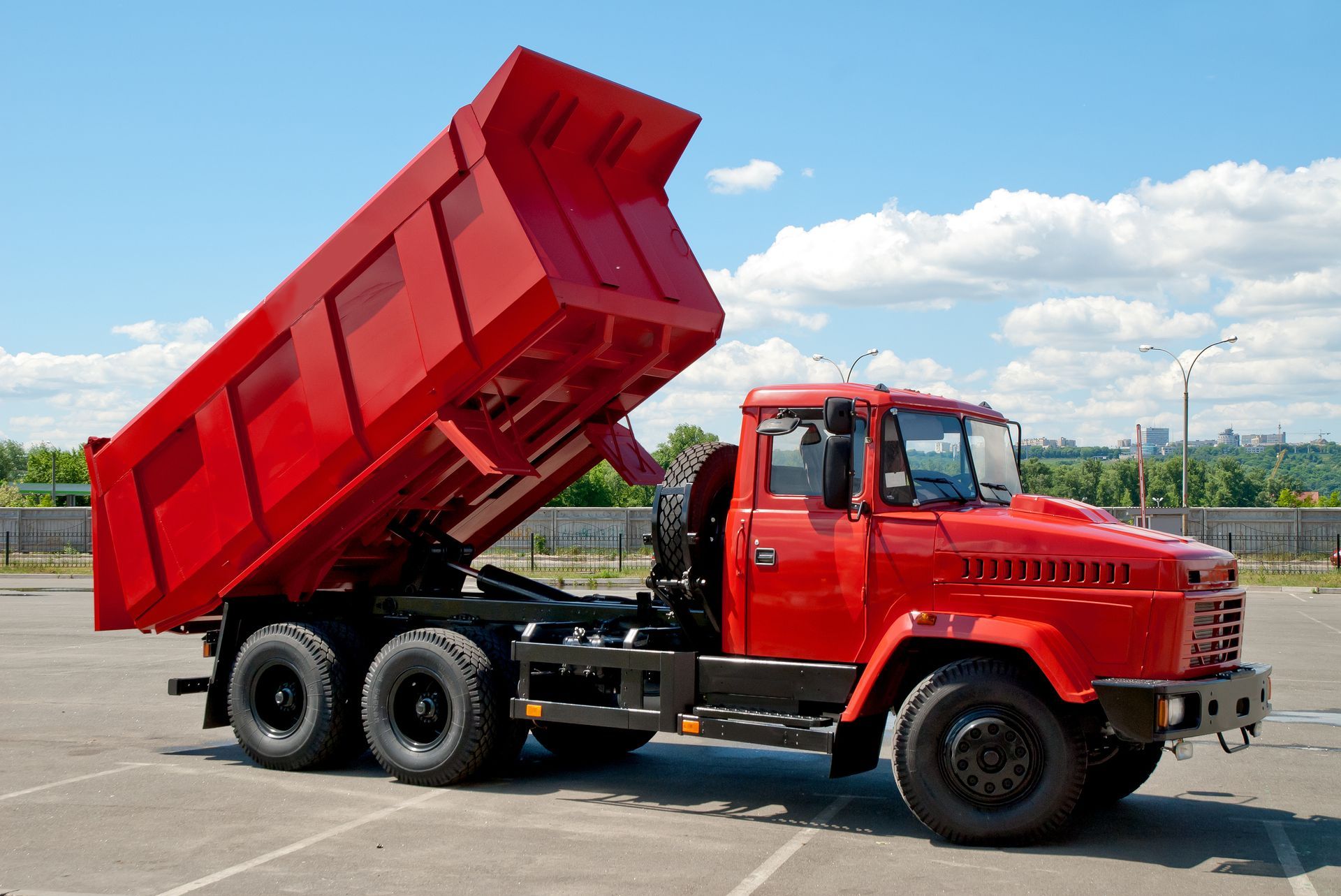 Red dump truck with raised bed parked on road under blue sky with clouds.