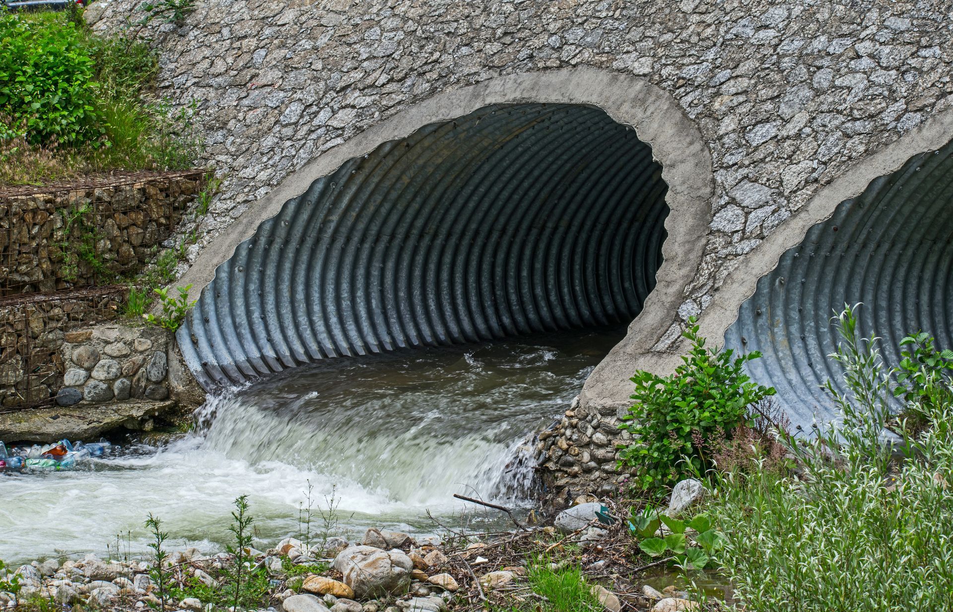 Two corrugated metal culverts under a stone wall with rushing water flowing through.