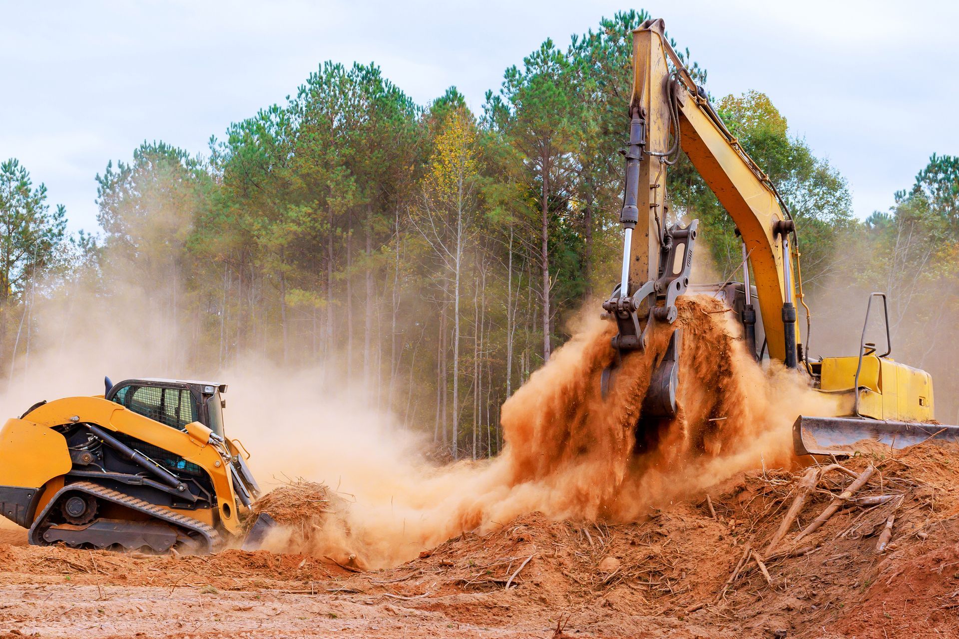 Yellow excavator and skid steer moving dirt on a construction site, trees in the background.