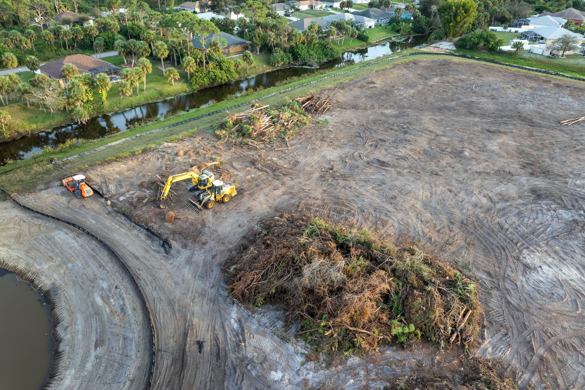 Excavator on a cleared construction site next to a canal, debris piles and a pond.