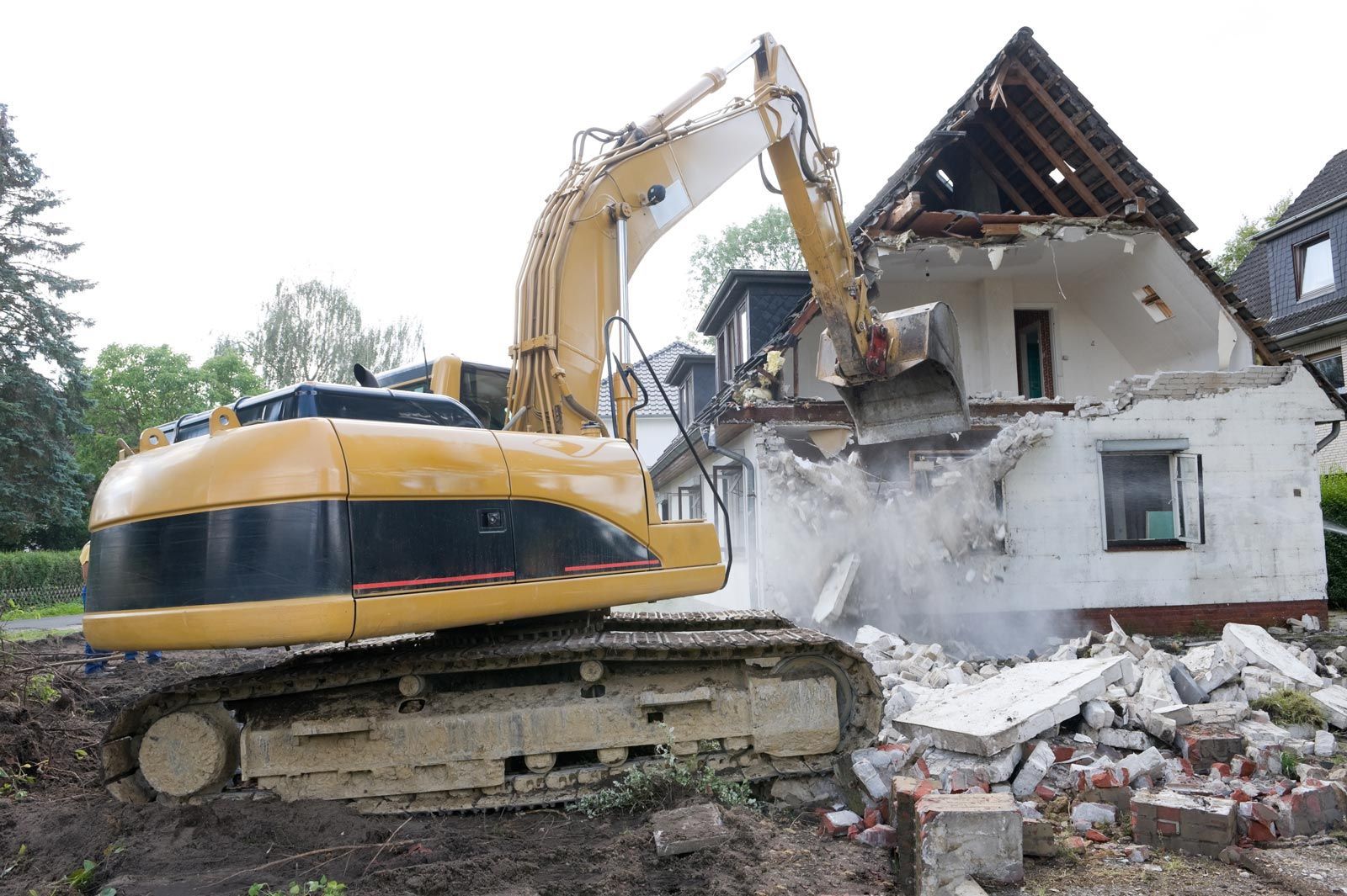 A large yellow excavator demolishes a white house; debris and dust surround.