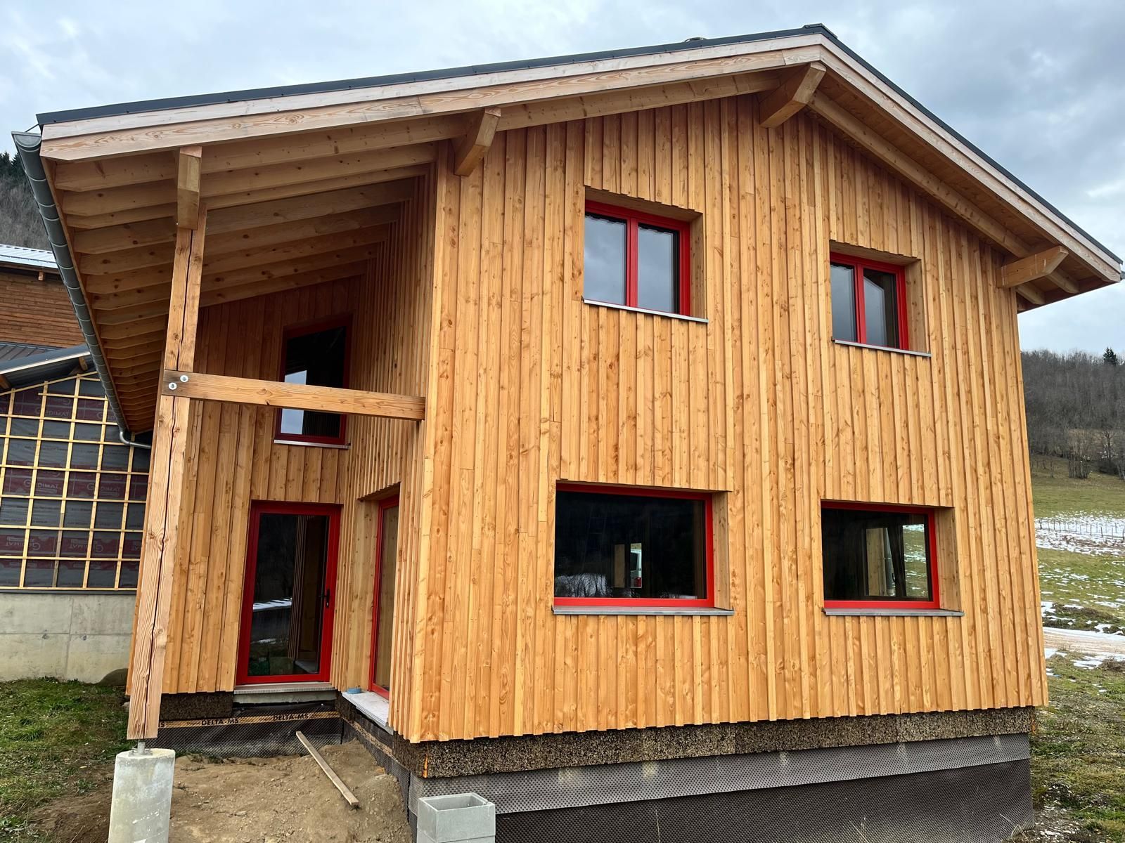 Wooden two-story house with red window frames, porch, and a partially built adjacent structure.