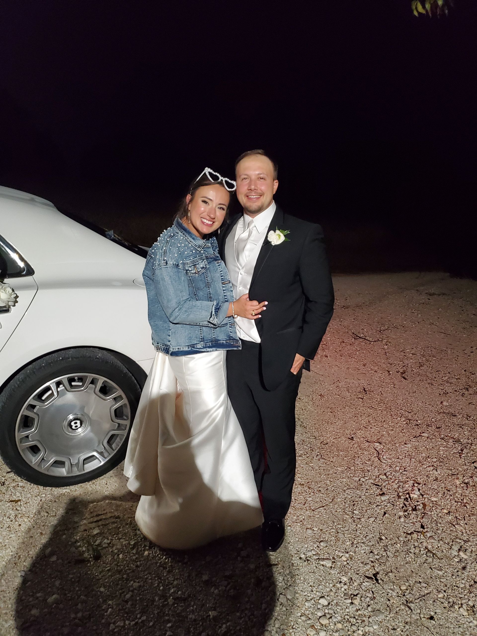 A couple in wedding attire, the person on the left wearing a denim jacket, standing outside at night next to a car.