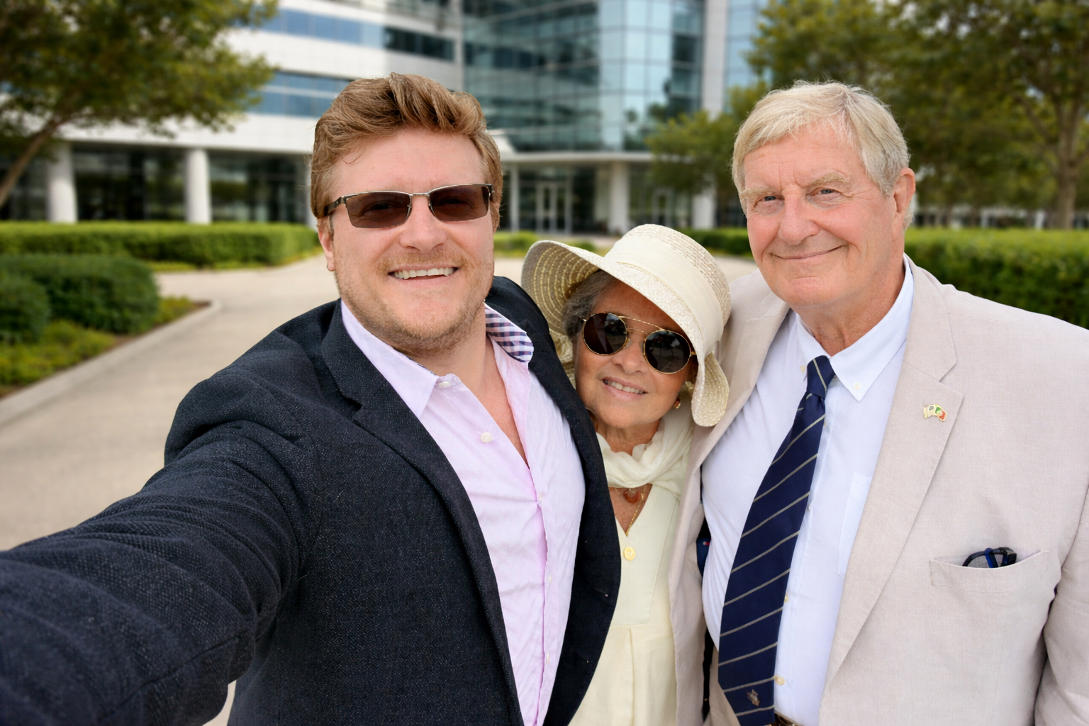 Three people smiling for a selfie in front of a modern glass office building with green hedges in the background.