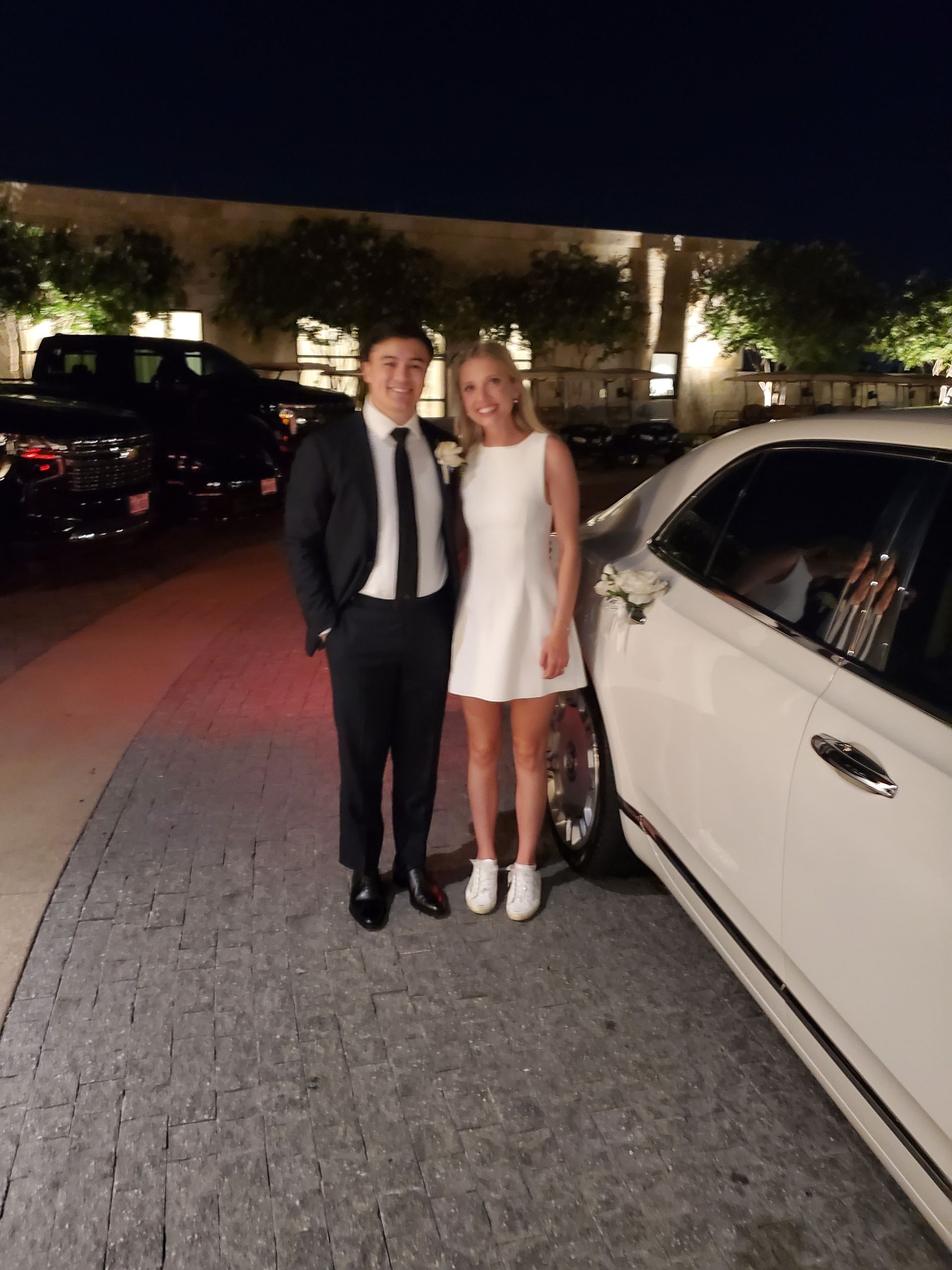 Man in suit and woman in white dress pose beside a white limousine at night.