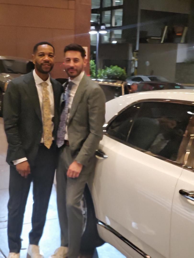 Two men in suits stand beside a white car. One has a gold tie, the other a patterned tie.