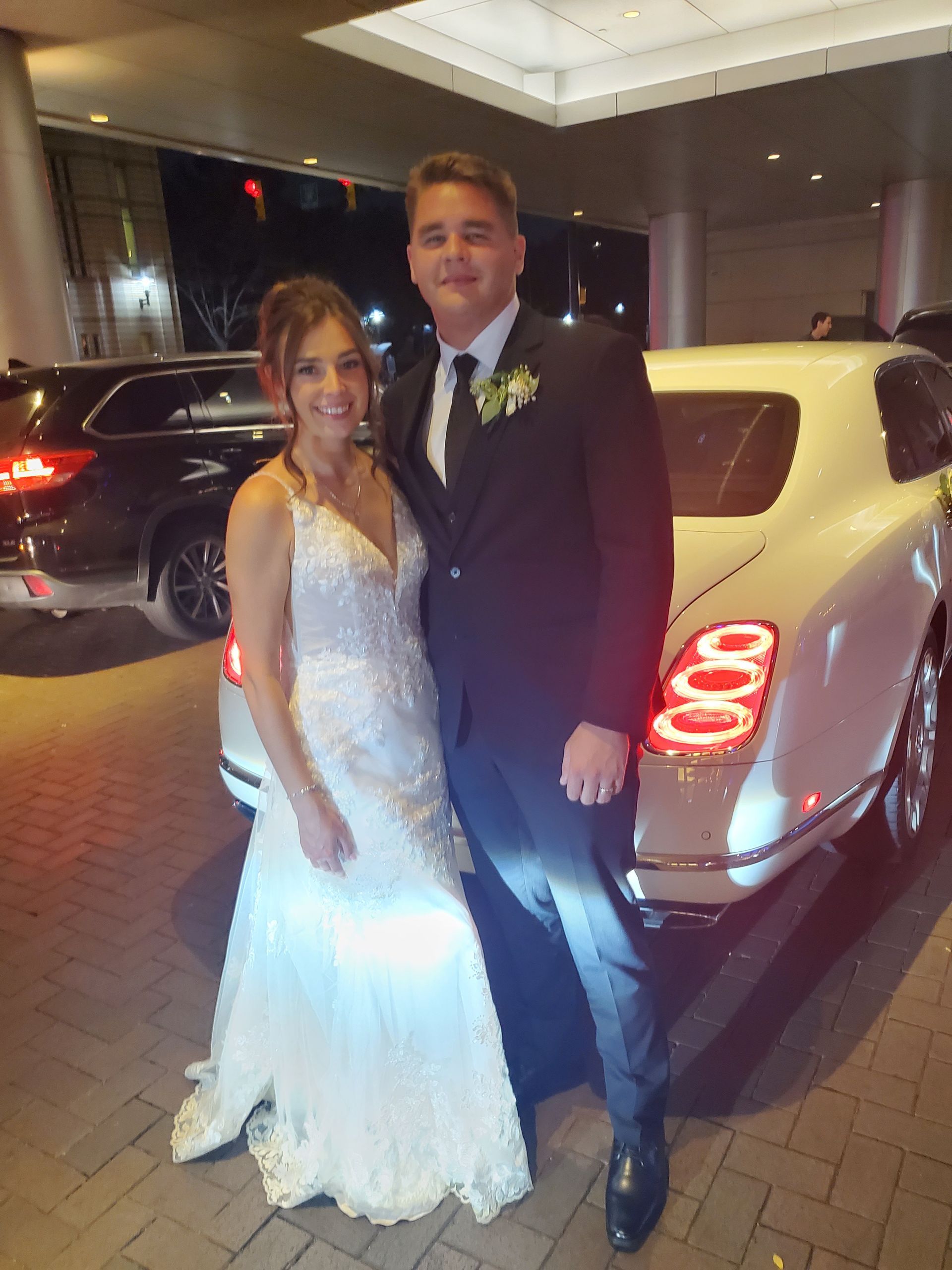 Couple in wedding attire posing by a white luxury car at night.
