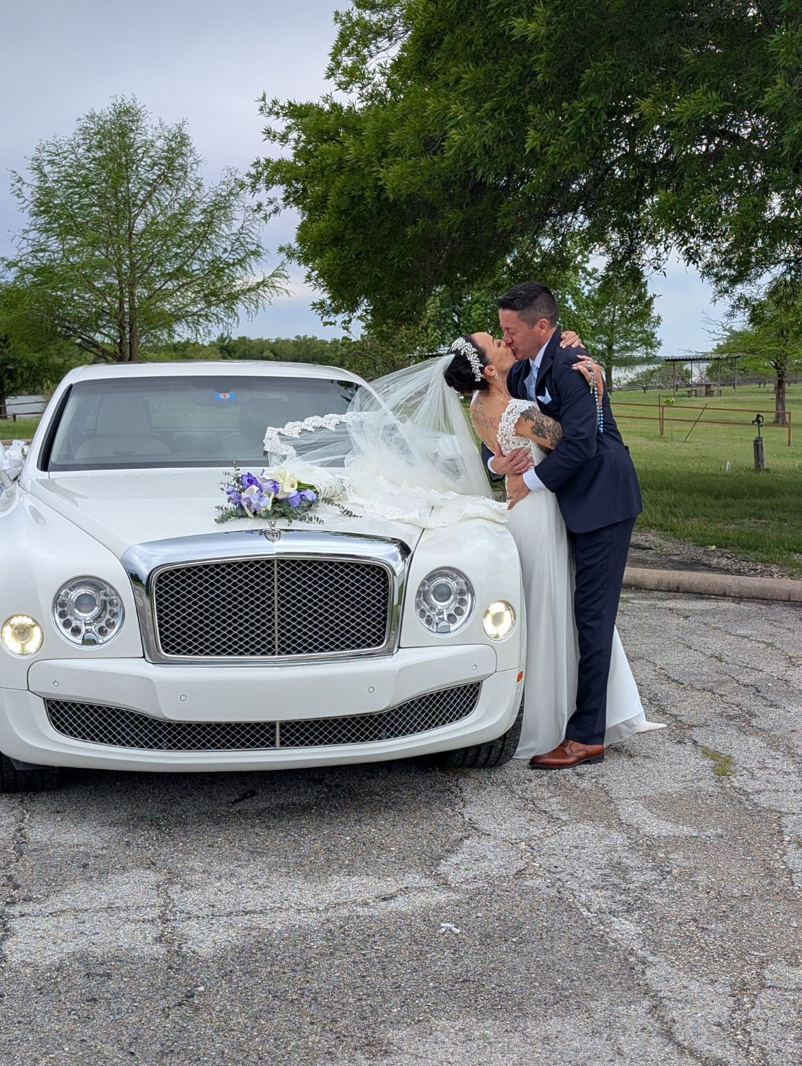 Bride and groom kissing beside a white luxury car with flowers on the hood outdoors