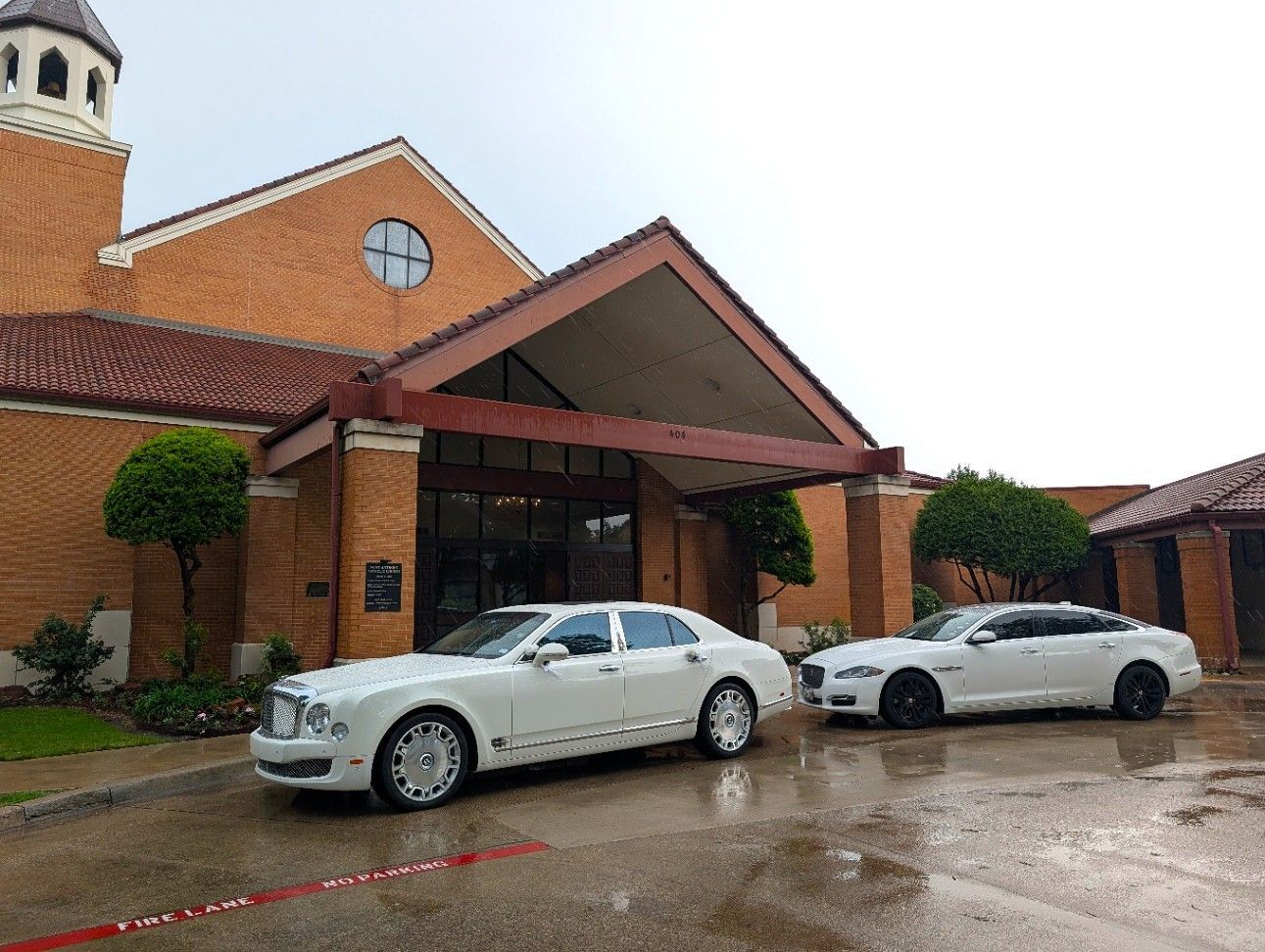 Brick building entrance with red roof and two white cars parked outside on a wet pavement.