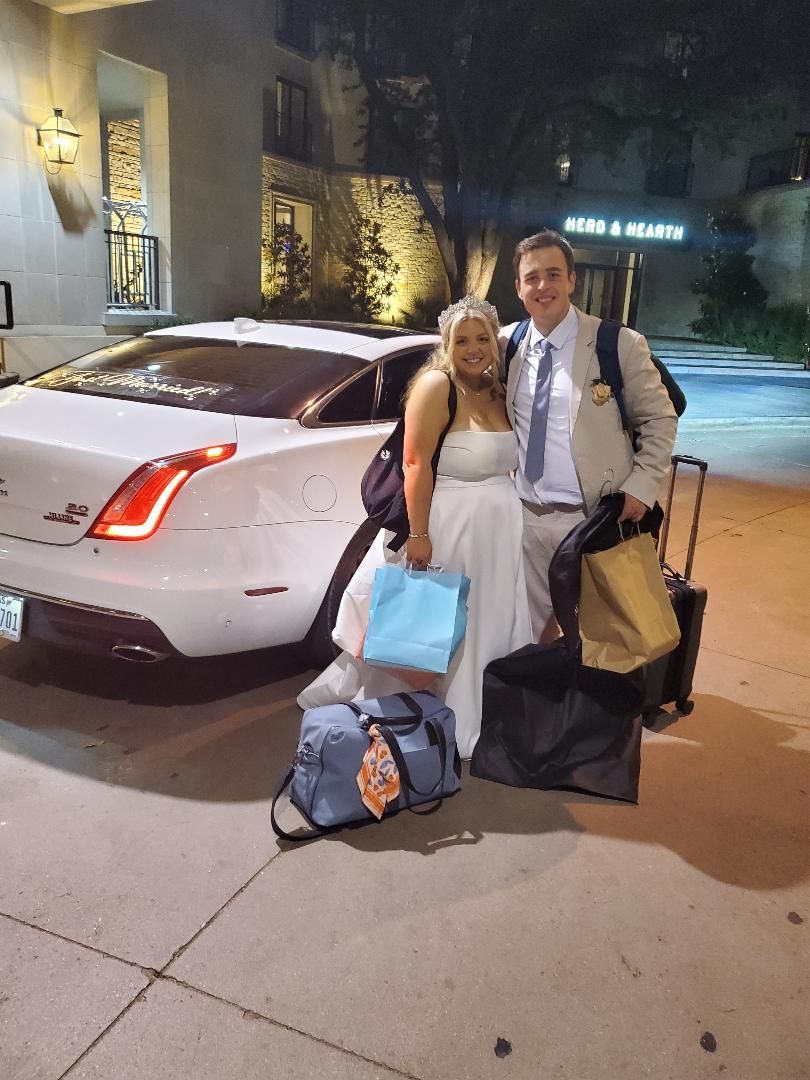 Newlyweds stand with luggage by a white car outside a building at night.
