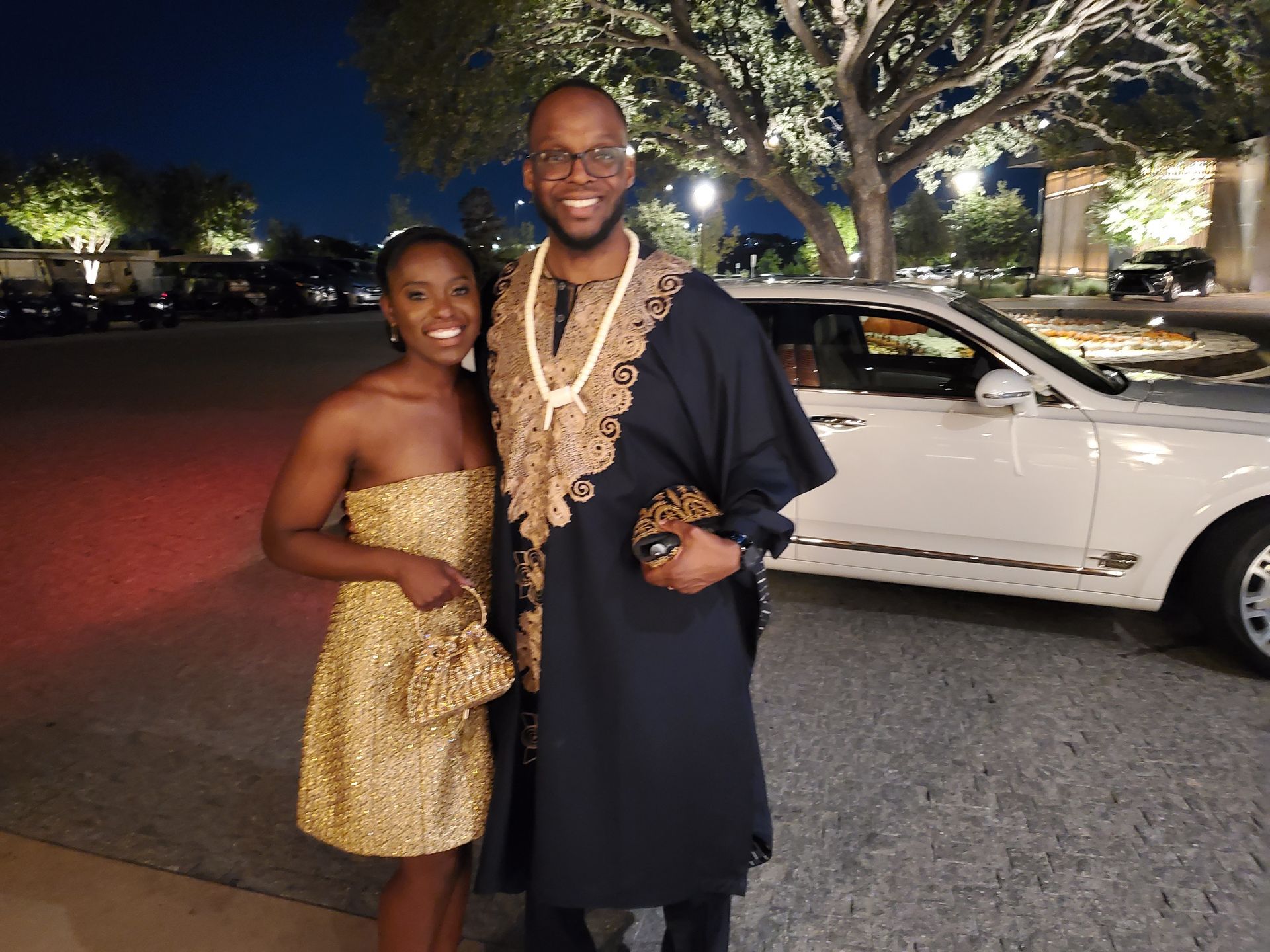 Couple posing by a white car at night. Woman in gold dress and purse; man in black and gold African attire.