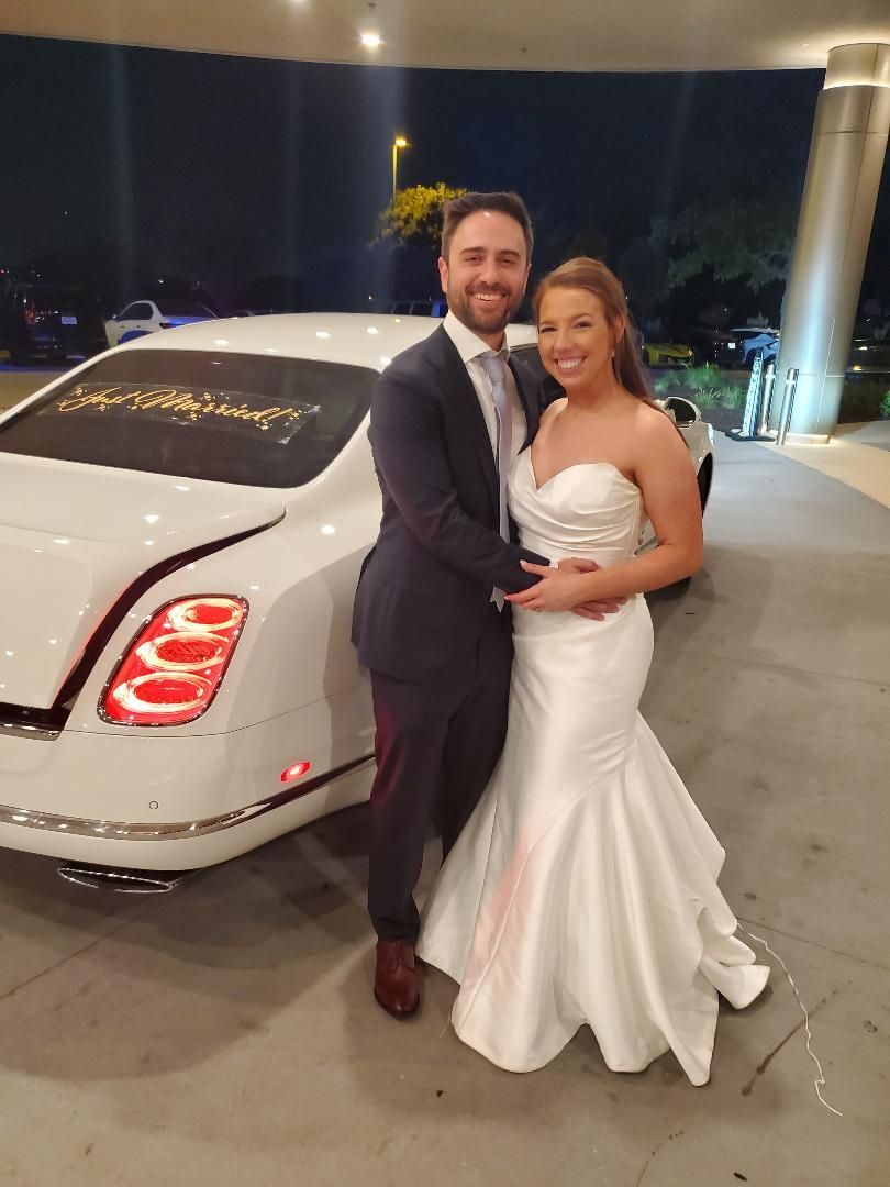 A newly married couple poses next to a white Bentley. The bride wears a white, strapless gown, and the groom a navy suit.
