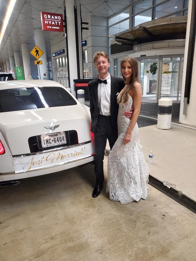 A couple in formal attire stands next to a white car with 
