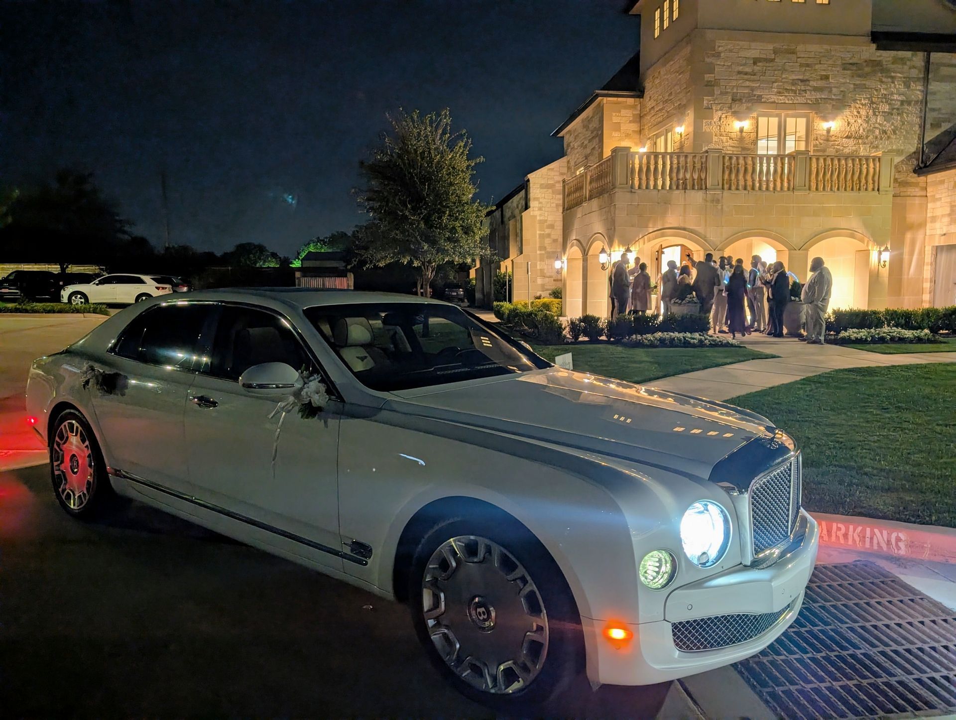White luxury sedan parked at night outside a brightly lit building with people gathered on the lawn.