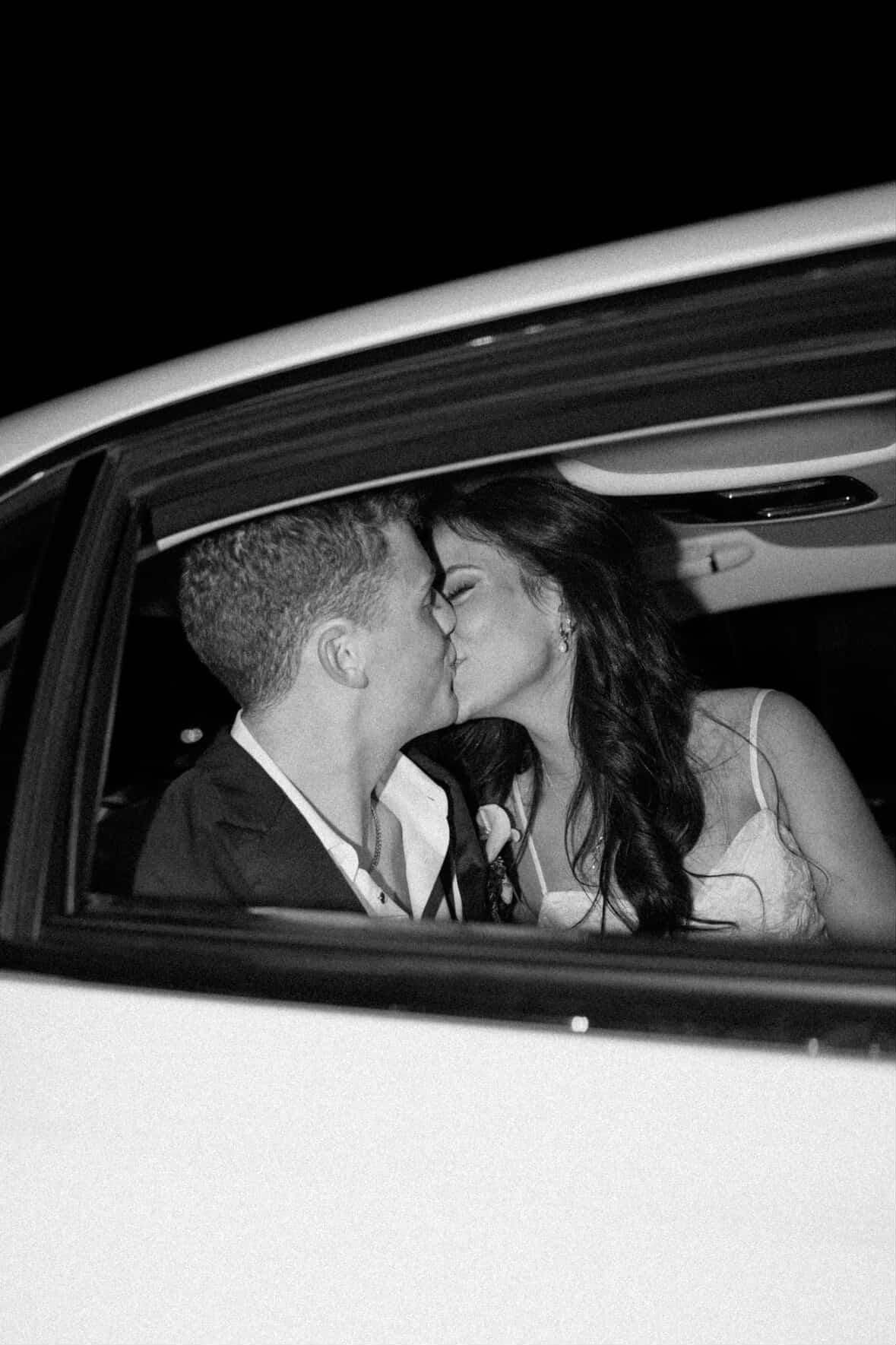 A black-and-white close-up of a couple kissing inside the back seat of a car at night.