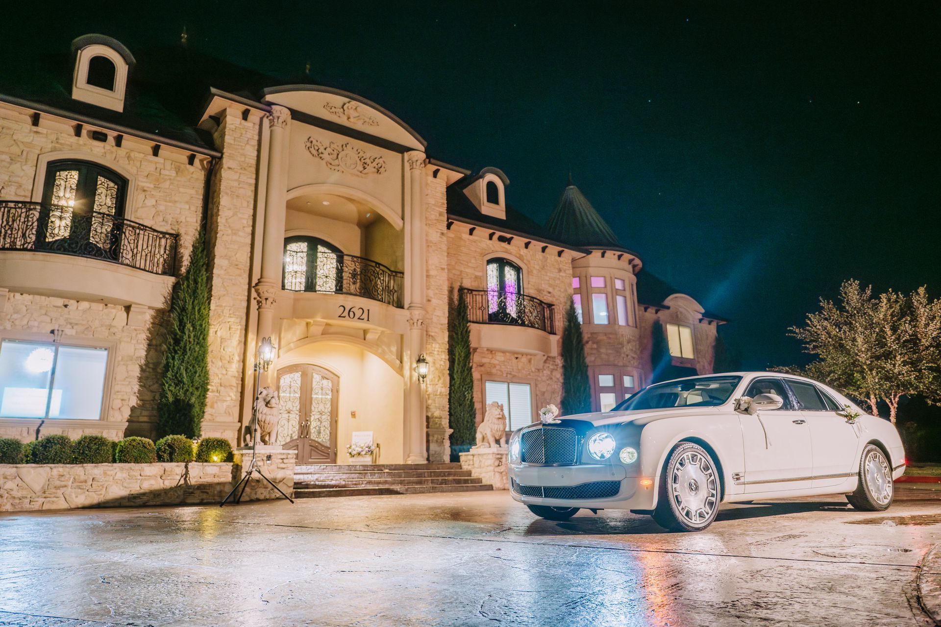 White luxury car parked in front of a large, lit mansion at night.