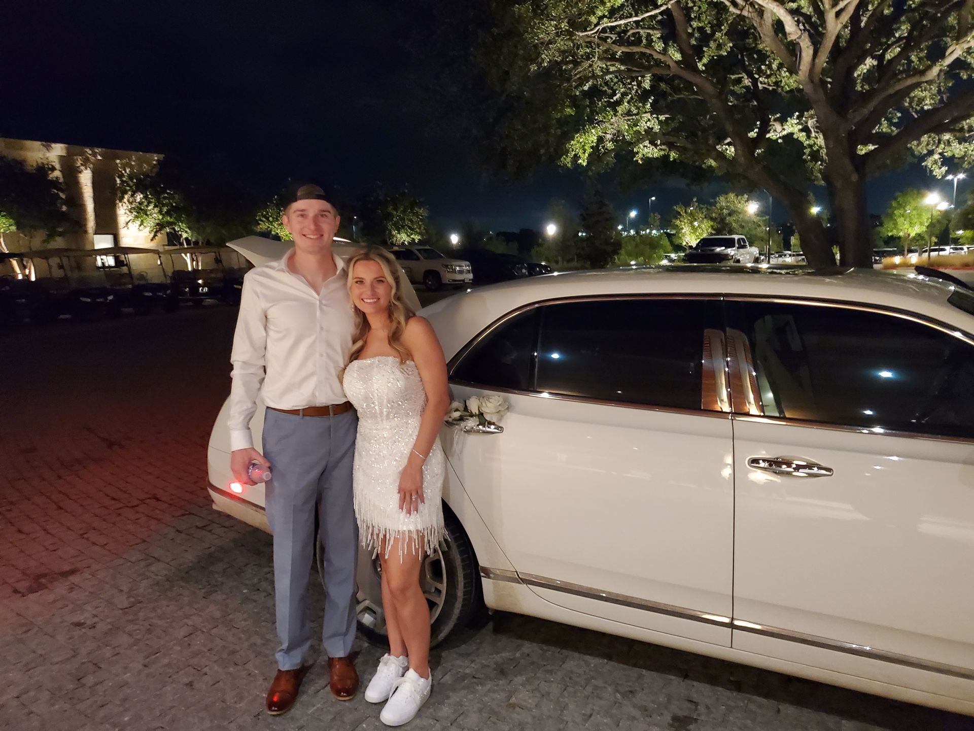 A couple poses by a white limousine at night. The woman wears a white dress and the man a button-down shirt and a cap.