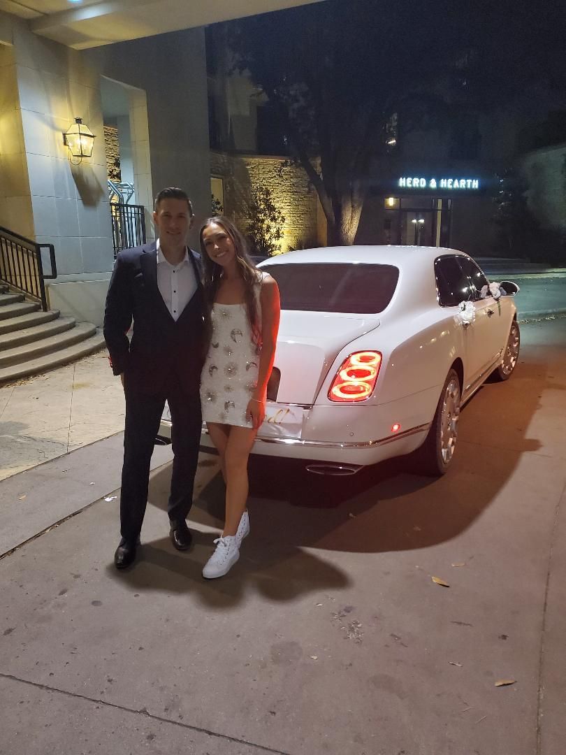 Man and woman posing next to a white luxury car at night. Man is in a suit, woman in a floral dress and sneakers.