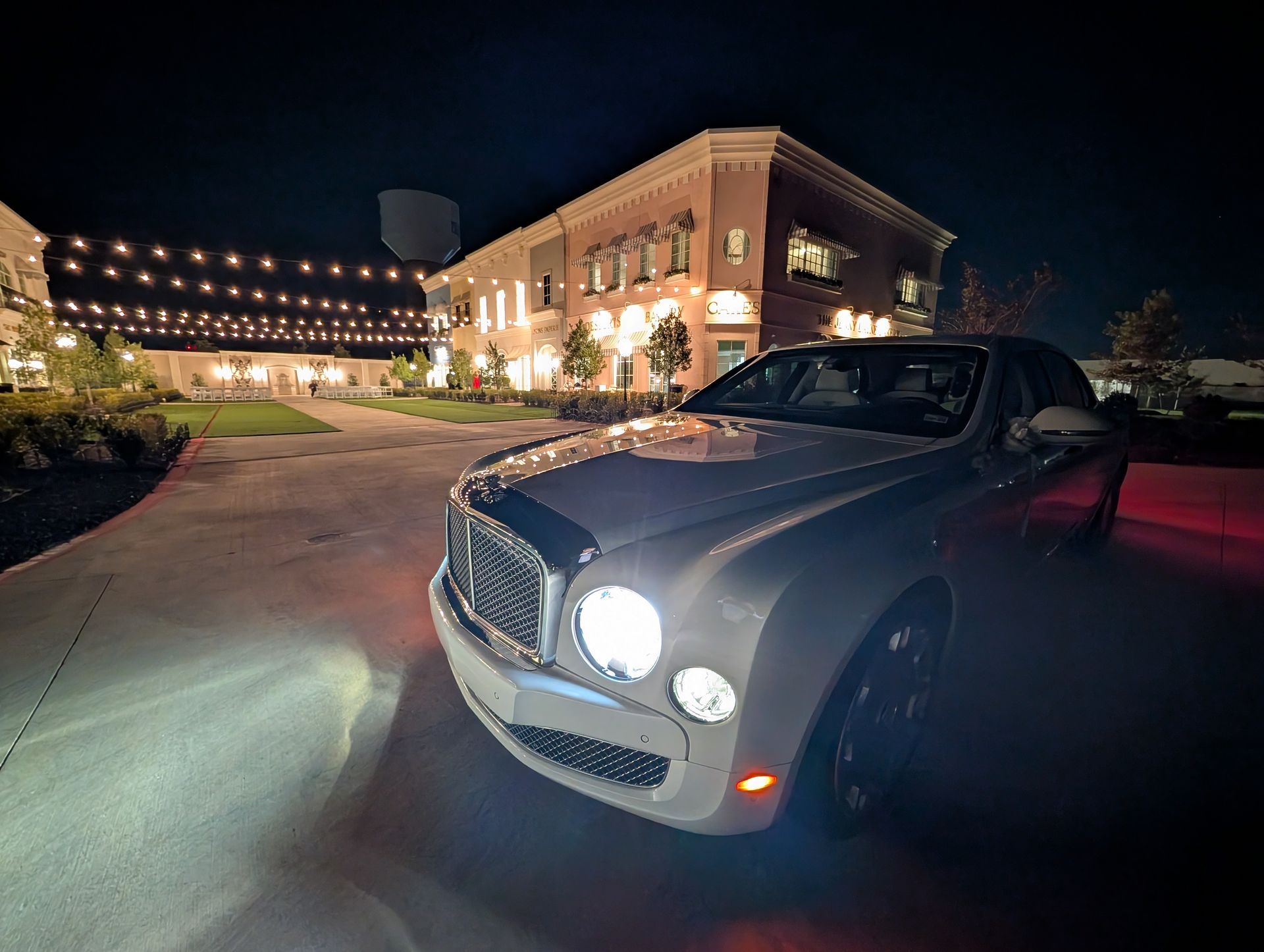 A silver Bentley parked in a lit courtyard at night in front of a multi-story building decorated with string lights.