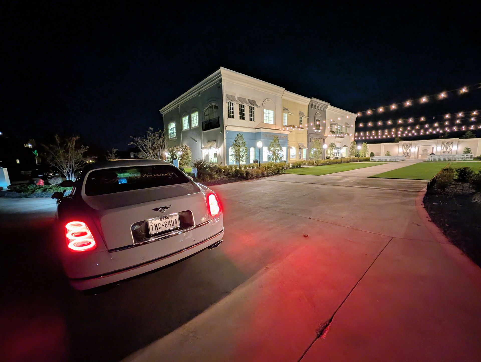 A white car parked in a driveway at night in front of a brightly lit, multistory building decorated with string lights.
