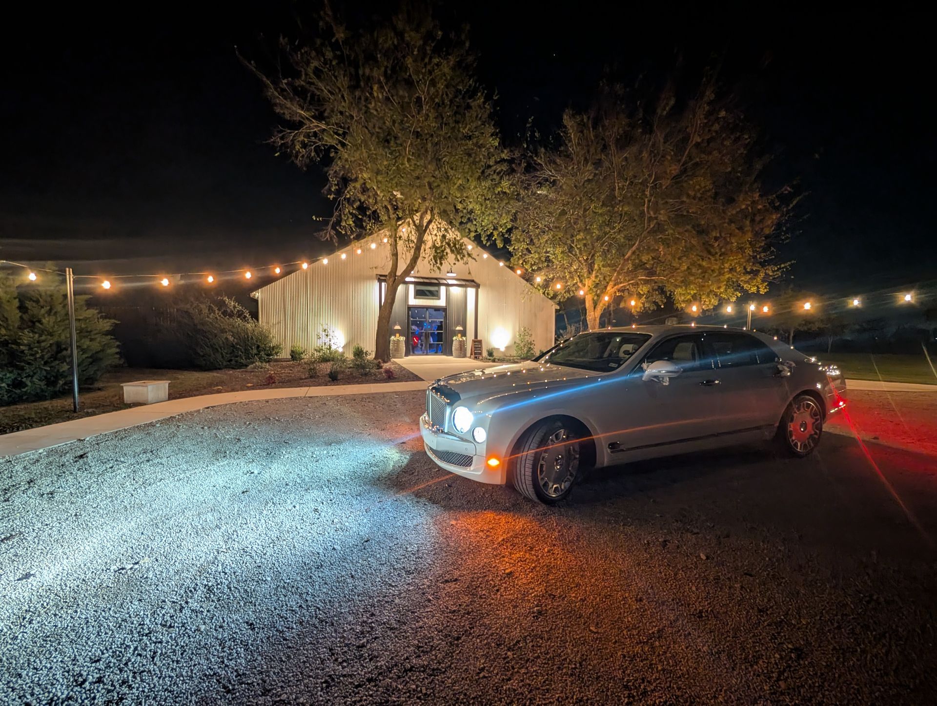 A silver luxury sedan with its headlights on is parked on a gravel lot at night in front of a lit, white barn venue.