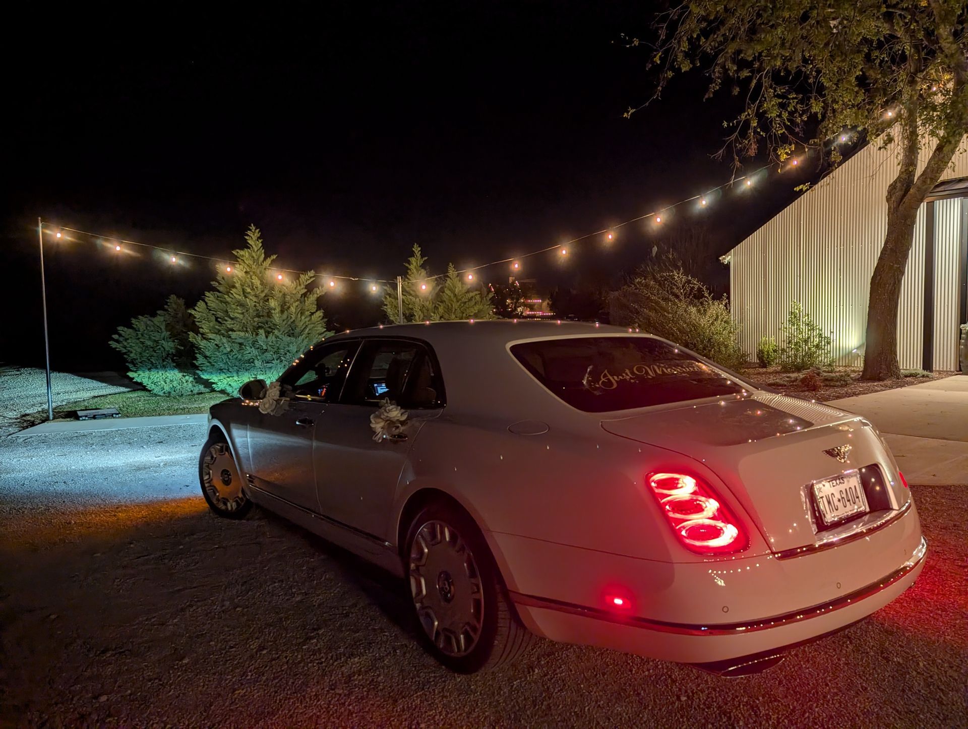 A white luxury sedan parked on a gravel lot at night, illuminated by string lights next to a white barn.