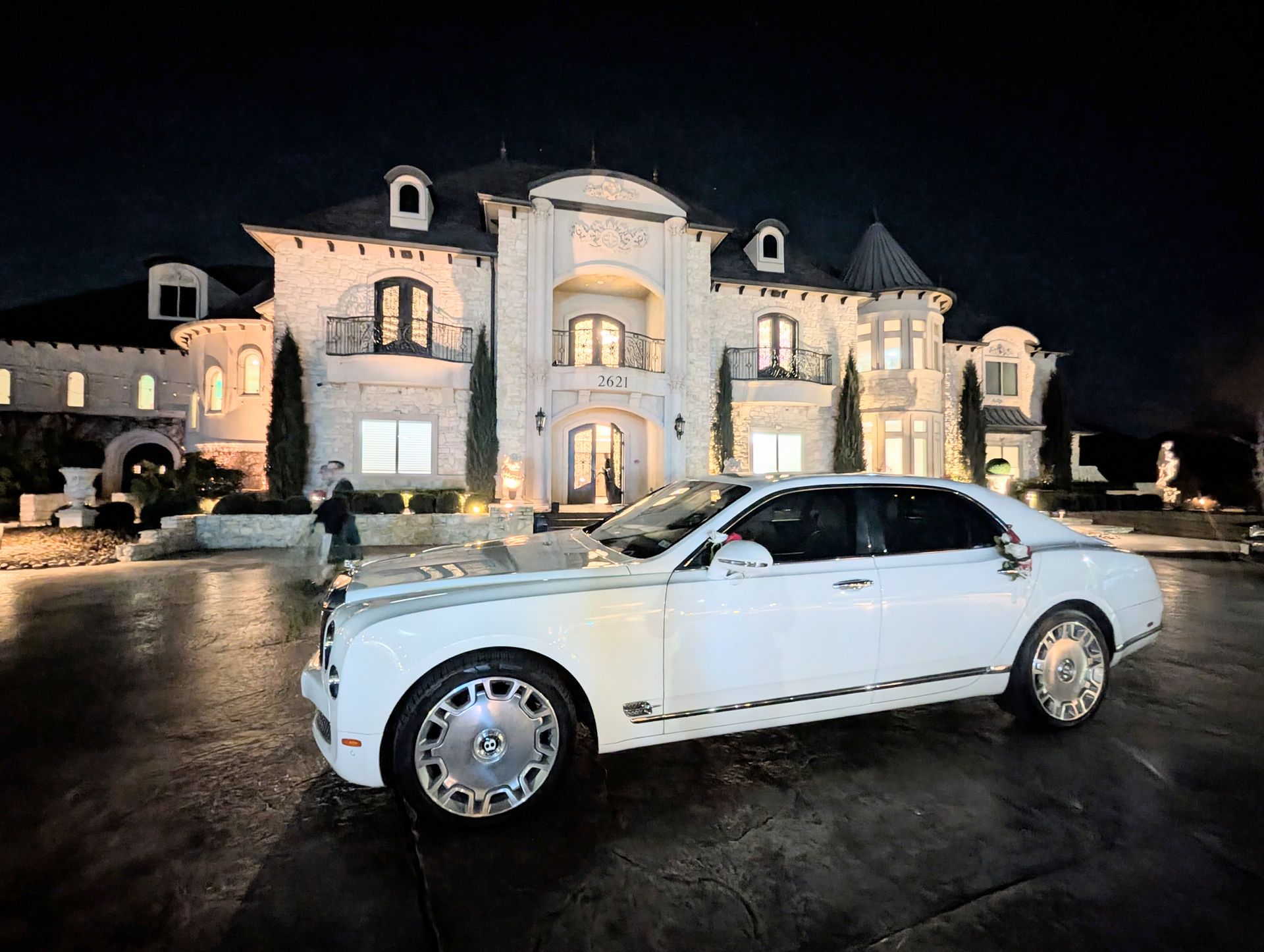 A white Rolls-Royce parked at night in front of a large, lit mansion with multiple windows and towers.