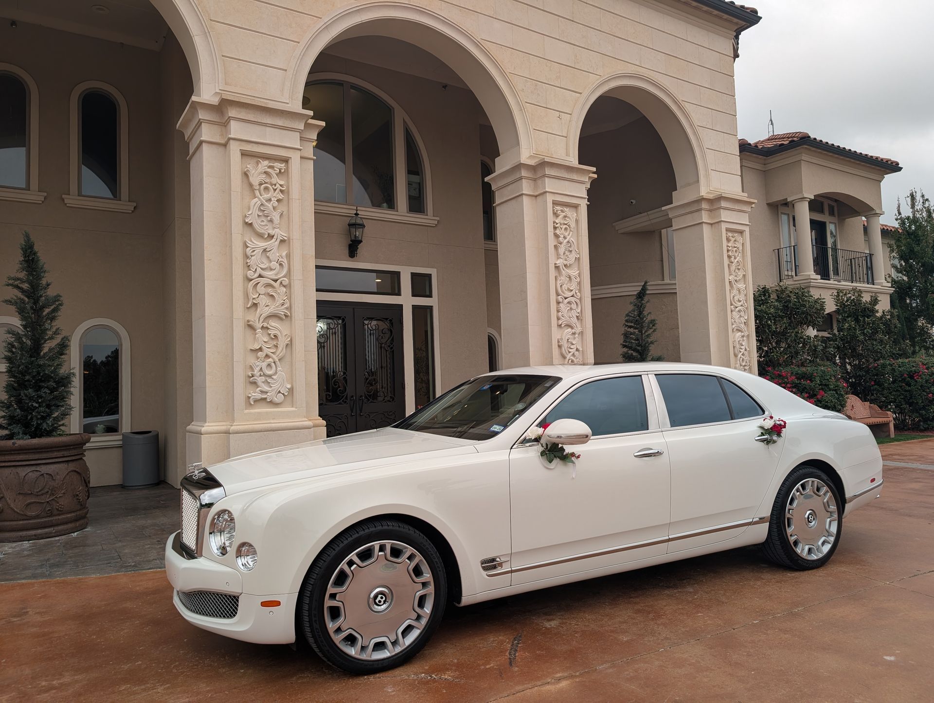 A white Bentley luxury sedan parked in front of a grand, tan stone mansion with arched entryways.