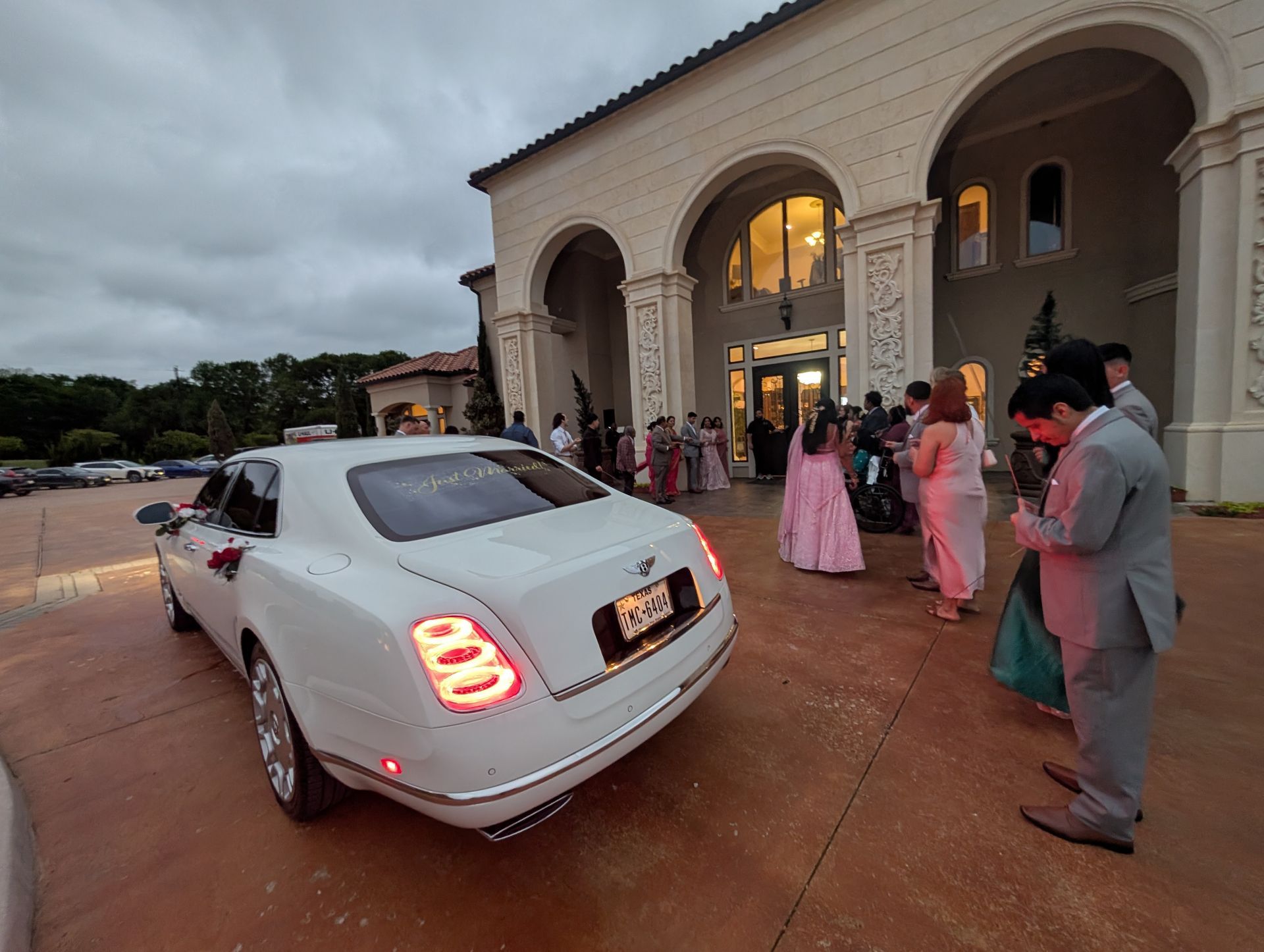 A white luxury car parked in front of a grand building entrance where guests in formal attire are gathered.