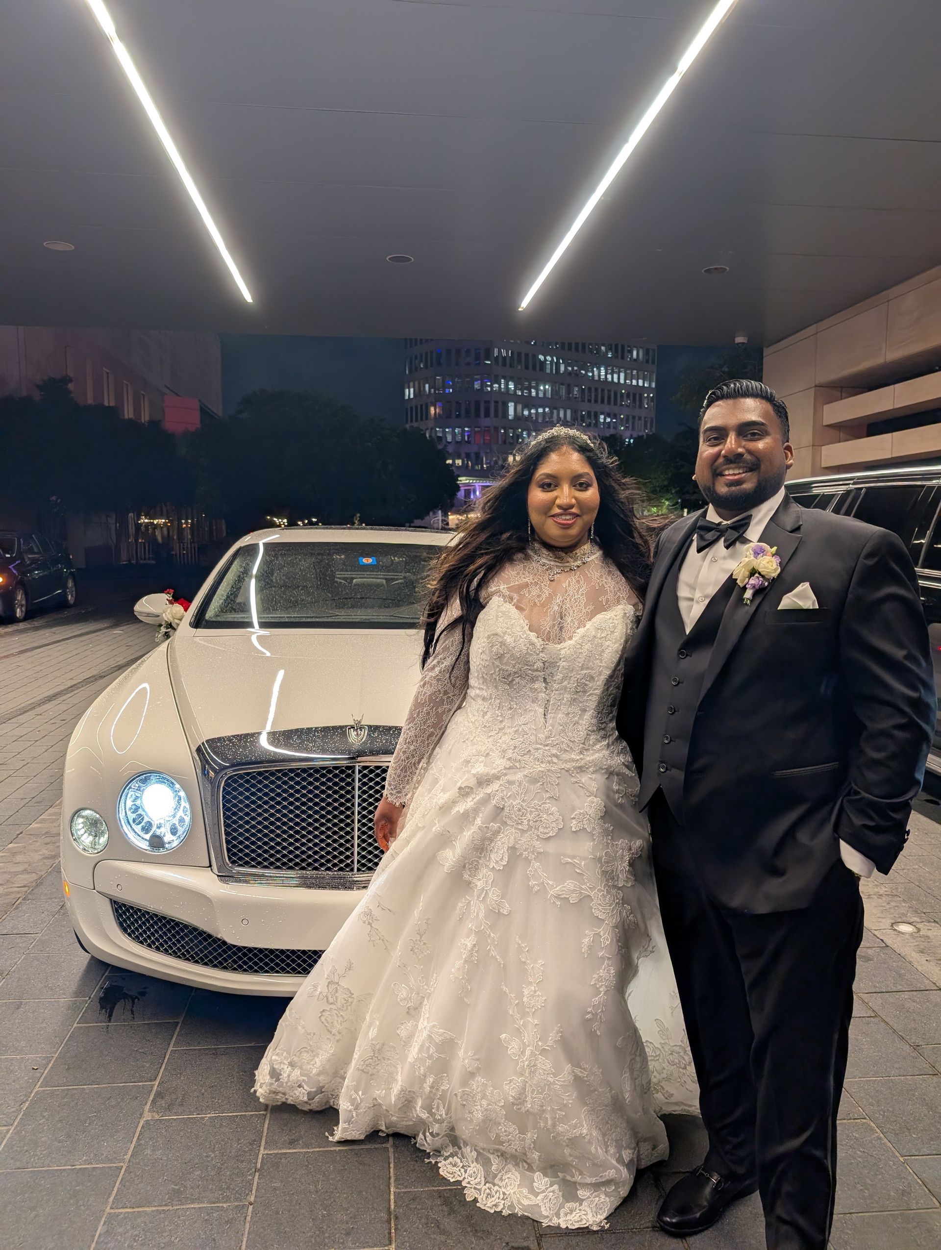 A smiling couple in formal wedding attire stands together in front of a white luxury car at night.