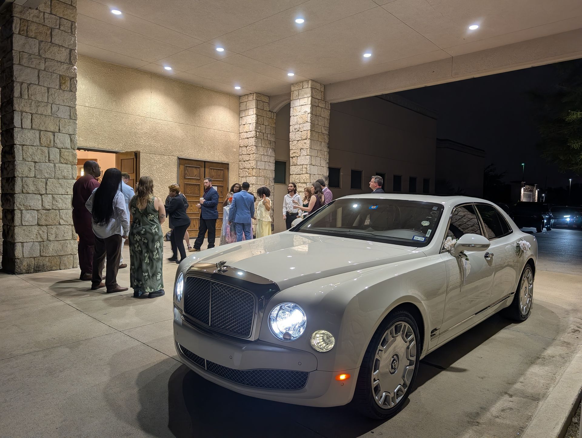 White luxury sedan parked under a hotel canopy at night, with people standing nearby.