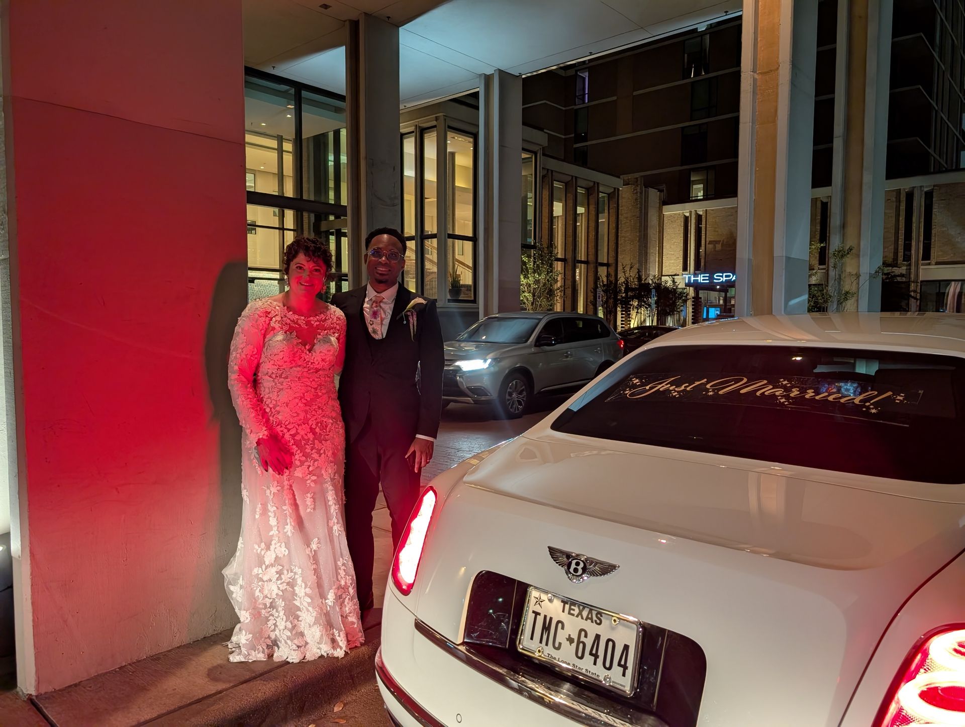 Two people posing beside a white luxury car at night under red lighting