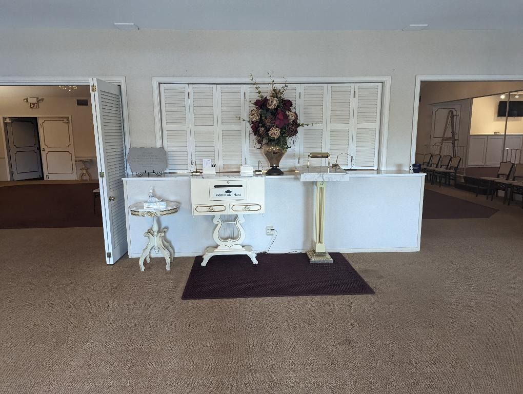 Reception area with table, floral arrangement, and doorways. White backdrop, brown carpet.