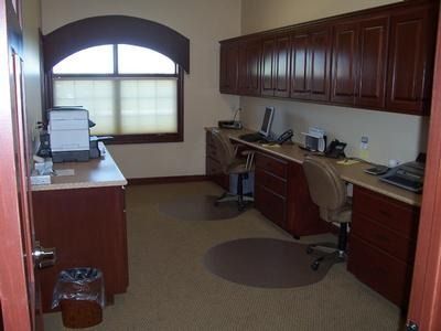 Office with two desks, computers, printer, cabinets, and a window. Brown wood furniture, beige walls, and carpet.