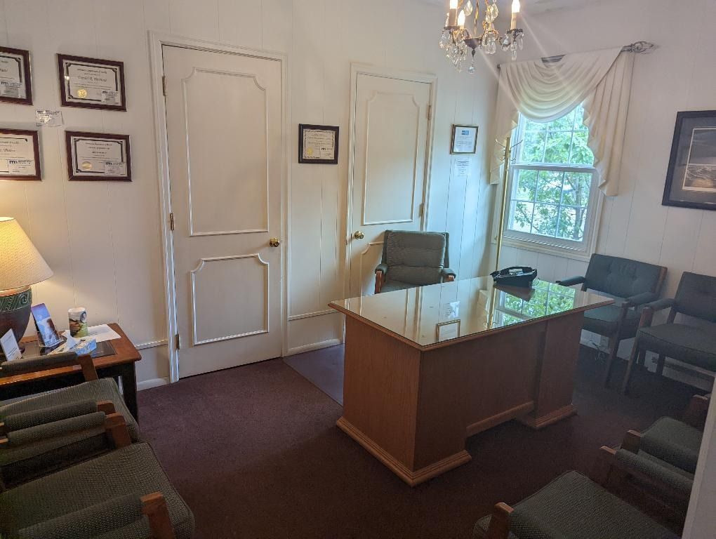 Office with desk, chairs, closed doors, and a window. Brown desk, green chairs, diplomas on wall.
